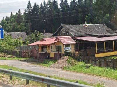 In Krestzy, Russia, a small, wooden kiosk with a red roof stands by a road. The kiosk has yellow siding and windows, one of which displays the word "ПИРОГИ" (Pirogi) in blue lettering. A pile of dirt and grass sits in front of the kiosk. Behind it, a weathered wooden fence encloses a property with several wooden structures, including what appears to be a covered outdoor area. Trees and a forest line the background, and a blue directional sign is visible to the left of the kiosk. A metal guardrail runs along the foreground, indicating the view is from a vehicle. The scene appears to be a rural roadside establishment.