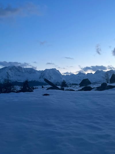 The image presents a winter landscape in Kovačka Dolina, Montenegro. The foreground is composed of an uninterrupted expanse of undulating snow, exhibiting a blue-white coloration. Several dark, irregular objects, consistent with rocks or partially buried debris, are visible within this snow layer. The midground features sparse, dark, bare shrubbery on the left, a large, dark, diagonally oriented timber or fallen log, and a solitary evergreen tree. Further into the midground, two dark-roofed, rustic structures, resembling cabins or barns, are situated on snow-covered terrain, with faint indications of fencing lines in their vicinity. The background is dominated by a continuous range of snow-capped mountains stretching across the horizon, displaying distinct peaks and contours. The sky above is a gradient of deep blue, lightening towards the mountain line, with scattered, thin, dark-grey clouds present. No individuals, animals, or discernable activities are present in the scene.