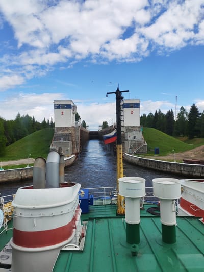 A ship is traversing the Volga–Baltic Waterway in Анхимово, Russia, approaching Lock Number 2. The lock is flanked by identical white buildings, each marked with a large red "2". A Russian flag is visible on the vessel. The sky is bright blue with scattered cumulus clouds. Green grassy slopes and trees border the waterway. The foreground shows the ship's deck, featuring white and red structures, green deck paint, and two white vents with green bases.