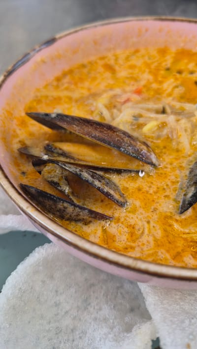 A close-up, eye-level shot shows a pink-rimmed bowl filled with a creamy orange soup. The soup contains strands of noodles and several mussel shells, some of which are partially submerged. The mussels are dark brown and appear to be cooked. The bowl rests on a textured white surface, possibly a napkin or placemat, and a blurry grey surface is visible in the background. The lighting is natural and illuminates the soup's rich color and texture. The image appears to be a food photograph taken in the city of Zarasai, Lithuania, likely in a restaurant or home setting.