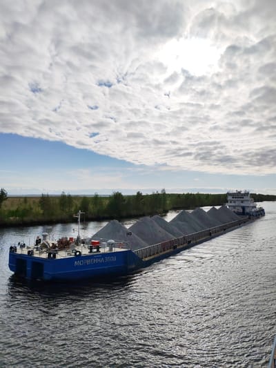 The image shows a large barge laden with piles of grey gravel or sand, moving along a waterway near Ozernoe Ust'e, Russia. The barge, with "MOPBEHHA 3608" painted on its side, is being pushed by a tugboat visible in the background. The waterway is flanked by a line of green trees and vegetation on either side. The sky is partially cloudy, with the sun shining through a break in the clouds. The water is rippled.