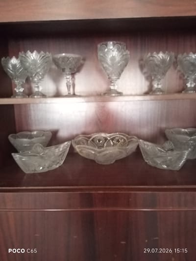 The image shows a wooden cabinet with two shelves filled with various crystal glassware. The upper shelf displays eight individual stemmed glasses, each with intricate cuts and designs. The lower shelf holds several shallow bowls and serving dishes, also made of cut crystal. The lighting in the image is dim, suggesting an indoor setting. The cabinet itself appears to be made of dark wood with a polished finish. The bottom right corner of the image contains a timestamp indicating "29.07.2026 15:15" and "POCO C65". This image was taken inside a cabinet in Beruniy, Uzbekistan.