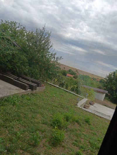 The image depicts an outdoor scene, possibly a backyard or a rural property, with a view of a small town or village in the background. The foreground is dominated by a patch of uneven green grass with scattered weeds and some dry patches. To the left, there is a concrete slab with stacks of dark, rectangular objects, possibly construction materials like lumber or metal beams. Adjacent to the slab is a dense green bush or tree. Further into the scene, a path made of concrete slabs leads towards a small, light-colored building with a dark roof. Two outdoor lamps are visible along the path. In the mid-ground and background, there are numerous trees and bushes, interspersed with houses featuring red tile roofs, suggesting a residential area. The terrain appears to slope upwards towards the horizon. The sky is overcast with gray clouds, indicating a cloudy day. The image appears to be taken from an elevated position, possibly from a window or balcony, with a screen or netting partially obscuring the view. The location is identified as Suhache, Bulgaria.