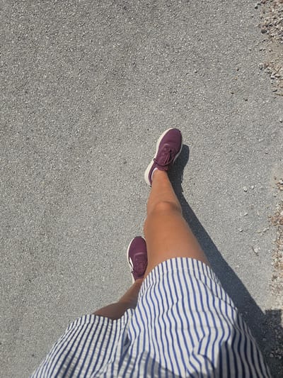 This is a top-down, close-up shot of a person's legs and feet, standing on a paved surface that appears to be asphalt with small pebbles. The person is wearing dark purple athletic shoes with white soles and laces, and a blue and white striped skirt or dress. A shadow, cast by an unseen object, stretches diagonally across the image from the upper right to the lower left, suggesting it's a sunny day. The scene is outdoors, likely in a casual setting like a park, walkway, or street. There is no visible text in the image. The lighting suggests it is daytime, and the clear shadow implies good weather. The focus is on the lower body of the individual, with the texture of the ground and the clothing being prominent. The specific location of Islamorada, USA, is provided as context, but there are no visual cues within the image itself to confirm this location. The overall mood is neutral and observational.
