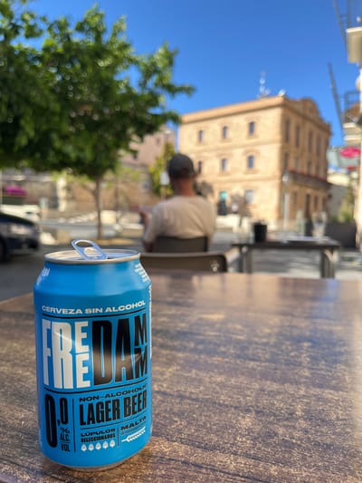 The image shows a can of Freeday 0.0% Non-Alcoholic Lager Beer, placed on a table, likely at an outdoor cafe. The label reads “CERVEZA SIN ALCOHOL” (beer without alcohol) and “LAGER BEER”. The pull tab on the can is visible.
In the background, there is a blurred view of a tree and a multi-story building with multiple windows. A person wearing a cap and light-colored shirt is seated at a table. The scene appears to be outdoors on a sunny day in Calafell, Spain.