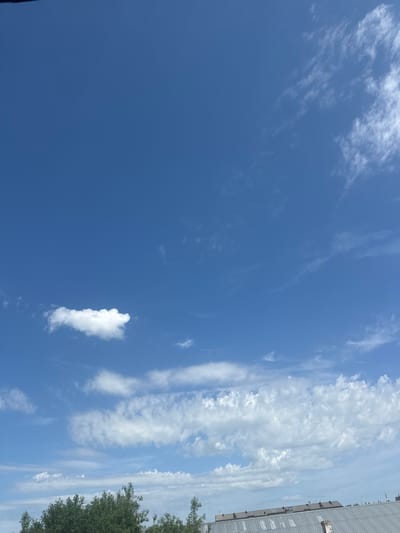 The image captures a wide expanse of a bright blue sky with scattered white clouds. In the lower portion of the frame, the tops of green trees and a metallic roof are visible, suggesting an urban or suburban setting. The scene appears to be a clear day in Aqköl, Kazakhstan, with no people or specific actions depicted. The focus is on the natural elements of the sky and vegetation, with hints of man-made structures at the bottom.