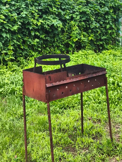 A rusty, portable barbecue grill stands on four thin legs in a grassy area. The grill is made of dark red metal and has a rectangular firebox with ventilation holes along the sides. A black metal ring, possibly for supporting cookware, is affixed to a central support within the grill. Behind the grill, a dense wall of vibrant green foliage forms the background, with the lower portion of the foliage transitioning into a lower-lying, bright green ground cover. The scene appears to be outdoors in a natural or semi-natural setting, likely in an urban area given the context of its location in Konstantinivka, Russia. No people are present in the image, and no actions are being performed.