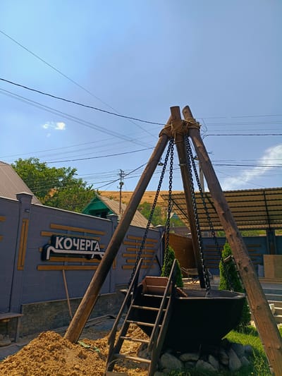 In the city of Kok Jar, Kyrgyzstan, a low-angle outdoor shot captures a unique structure with a rustic aesthetic. Three large wooden logs are propped up at an angle, bound together at the top with thick rope. Hanging from this structure are several heavy chains, which support a large, dark basin. A wooden ladder leans against the basin, with one step visible at the top. In the background, a grey brick wall displays the word "КОЧЕРГА" in large white letters with a black outline. Behind the wall, a green building and trees are visible, with a dry, grassy hill in the distance. Power lines crisscross the clear blue sky. The ground in the foreground is covered with a pile of light brown soil.