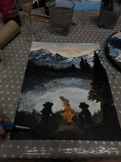 An indoor scene depicts a painting session in progress, laid out on a grey tablecloth adorned with white star patterns. The central focus is a canvas painting, displayed vertically in the image but likely intended horizontally, portraying a dramatic landscape. The artwork features dark, silhouetted mountains and trees on the left, contrasting with a lighter sky or fog in the middle. On the right, two figures, possibly cowboys or Western-style individuals, are silhouetted against a bright, warm light source, likely a campfire, which adds orange and yellow hues to the dark foreground. The style is expressive with visible brushstrokes, primarily using black, white, grey, orange, and yellow.
Surrounding the painting are various items. A paint palette, holding blobs of blue, white, and grey paint, is visible at the top. Several paintbrushes are scattered, with one blue-handled brush actively held by a hand at the bottom right, poised over the canvas. Two clear drinking glasses, one containing a dark liquid, and a stemmed wine glass with a light-colored beverage, are positioned on the left. A partially visible red cylindrical container, identified by its text as a Coca-Cola product, is also on the table. The visible text on this container includes "Core Contains Source of," "Carbohydrate," "100ml 250ml Store conta," "0 Skcal 1kcal," "Coca-Cola Recycle," "2023 European," "I'm a bottle made fon 100," and "Try More Weird," along with "ACRIUL FANT WO CCEP GB UB8 1EZ 0808 227711." A brown cardboard tube, possibly a paper towel roll, completes the array of items. The setting suggests a casual and creative leisure activity, possibly an art class or a relaxed hobby session in Ipswich, United Kingdom.