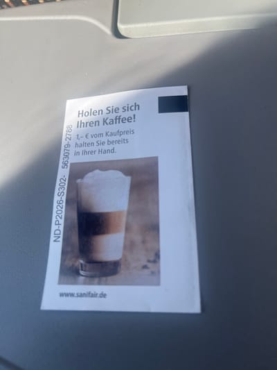 The image shows a close-up of a flyer on a grey surface, possibly a tray table or seat. The flyer is oriented vertically and features text and an image of a coffee drink. The text on the left side of the flyer is in German and reads: "Holen Sie sich Ihren Kaffee! 1,- € vom Kaufpreis halten Sie bereits in Ihrer Hand." This translates to "Get your coffee! You already have €1 off the purchase price in your hand." Below this, a product code "ND-P2026-S302-563079-2788" is visible, along with a website address "www.sanifair.de" on the right side. The image on the flyer depicts a layered coffee drink, likely a latte or cappuccino, in a clear glass. The lighting suggests it is daytime, and the context of the German text and the website implies the flyer is related to a service or establishment in Germany. The overall scene is simple, focusing on the promotional material.
