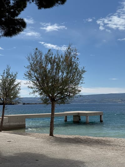 The image shows a concrete pier extending out into the Adriatic Sea in Dugi Rat, Croatia (latitude: 43.446, longitude: 16.636). The pier is made of concrete and has a square shape. The water is a clear blue and there are a few small waves. A tree is visible on the left side of the image and a large tree is located in the center of the image. The shore is sandy and there is a small amount of sand on the concrete pier. In the background, a mountain range is visible. The sky is blue and there are a few white clouds. The image was taken on a sunny day. There are no people in the image.
