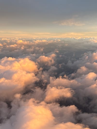 An aerial view presents a dense, undulating expanse of clouds under a transitioning sky. The cloud tops exhibit a warm, golden-orange and peach coloration, indicative of direct sunlight from a low angle. Deeper and shadowed sections of the cloud formations display cooler grey-blue and darker grey tones. The cloudscape extends across the frame, forming a textured, three-dimensional surface. Above the cloud horizon, the sky graduates from a light peach-grey hue to a muted grey-blue at the zenith. No people, objects, or ground features are visible. The scene is located over the city of Penafiel, Portugal.