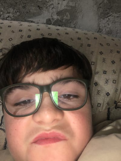 A close-up, upward-angled image depicts an individual, likely a juvenile, positioned in an indoor setting within the city of ქვედა სამება, Georgia. The subject has dark, short hair and is wearing dark-rimmed prescription glasses. Their eyes appear partially closed or looking downwards, with a distinct rectangular green reflection visible on the left lens. The lips are slightly parted, exhibiting a light pink color. The individual's facial expression conveys a subtle grimace or discomfort. Behind the subject's head is a beige or light-colored fabric with a repeating pattern of small dark squares and 'X' shapes, likely upholstery or a pillow. Above this fabric, a rough, grey, unfinished concrete surface is visible, suggesting a wall or ceiling. The lighting is direct, creating reflections on the glasses.