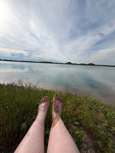 The image depicts a first-person perspective scene at the edge of a body of water in Sapunov Bor, Russia. In the immediate foreground, a pair of bare human legs and feet are extended forward. The left foot and lower leg appear largely clean with a few small dark specks, while the right foot and lower leg are heavily coated in a dark, wet, mud-like substance, extending up to the calf. The legs rest on or are positioned above a grassy bank, which features a mix of green and dry brown vegetation, along with areas of dry, cracked earth visible to the right.

Beyond the bank, a large, calm body of water occupies the midground, reflecting the cloudy sky above. The water's surface exhibits shades of light blue, grey, and some pale yellow, indicating its shallow depth or reflections. On the distant horizon across the water, a narrow strip of land is visible, featuring sparse trees, bushes, and several low-rise structures. The sky above is partially overcast with a mixture of white and light grey clouds, displaying both wispy formations and denser clusters, particularly towards the lower left horizon. The overall lighting suggests broad daylight.
