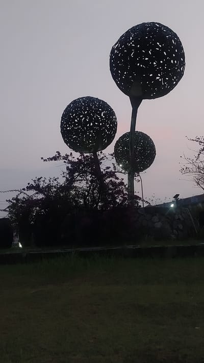 The image captures an outdoor scene at dusk, featuring three large, spherical sculptures with intricate, cutout designs, resembling stylized trees. The sculptures are dark in color and stand tall against a pale, twilight sky. In the foreground, a well-maintained grassy area is visible. Behind the sculptures, a dense cluster of dark foliage, possibly a shrub or tree with purple flowers, forms a dark mass. A low stone wall or embankment is partially visible behind the foliage, and beyond that, hints of urban structures, including lights and wires, can be seen. The overall atmosphere is serene, likely depicting a park or public garden space within Pusakarakyat, Indonesia, as indicated.