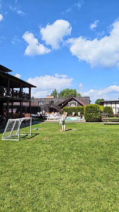 The image depicts a sunny day at an outdoor recreational facility. In the foreground, a well-maintained green lawn is visible, with a white soccer goal on the left and a blue soccer ball on the grass. A child, dressed in light-colored clothing, is standing in the middle of the lawn. In the background, there are several wooden buildings with multi-level structures and balconies, likely part of a resort or club. A swimming pool is situated behind a row of lounge chairs, and more seating is arranged around it. Green bushes and trees form a natural backdrop to the scene. The sky is a vibrant blue with scattered white clouds. The location is identified as being within the Занарацкі сельскі Савет, Belarus.