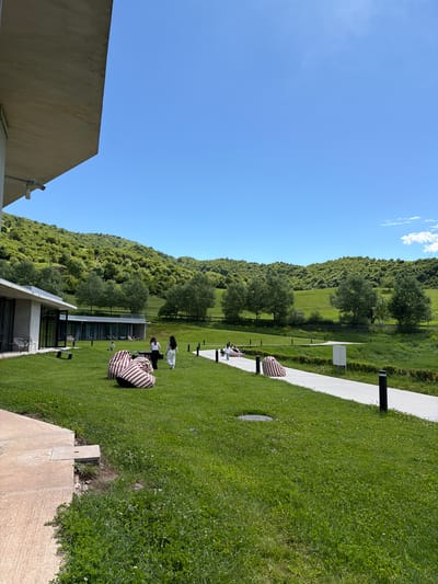 The image depicts an outdoor scene likely in Chkalov, Armenia. A grassy lawn extends from the lower right towards the center, dotted with striped beanbag chairs and some people who appear to be reclining. A white walkway runs along the right side, edged with what looks like a manicured hedge. Beyond the lawn, a hillside covered in trees and greenery rises towards the horizon. The sky is clear and blue, with a few wispy clouds near the top edge. A building is partially visible on the left side of the image, with architectural details indicating a modern or contemporary design.