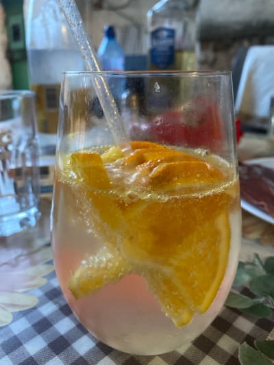 A close-up, eye-level, vertical shot captures a glass of sparkling water with orange slices and a straw. The drink is a light pink color and fizzes with carbonation, the orange slices are thin and appear fresh. A clear plastic straw with glitter is inserted into the drink. The glass sits on a checkered tablecloth. In the blurred background, other glasses and bottles are visible, suggesting a casual gathering or meal. The lighting is bright, highlighting the beverage. The location is identified as Miriantsi, Bulgaria.