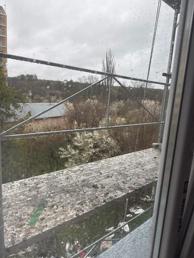 The image, taken from an elevated position, shows a view towards a forested area and buildings in Novi Pazar, Bulgaria. A metal scaffolding structure dominates the foreground, partially obscuring the scene. The focus is on the building under construction. Rain or condensation droplets are visible on the window through which the photo was taken, partially distorting the view. The sky is overcast.