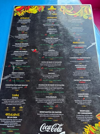 The image is a menu from a restaurant in Pozos, Costa Rica, located at the coordinates provided. The menu is printed on a white paper background with red and pink borders. The menu lists a variety of traditional Costa Rican dishes, including appetizers, soups, main courses, and desserts. There are also a few American-style dishes, such as hamburgers and tacos. The prices for each item are listed in Costa Rican colones. The menu is adorned with a stylized image of a chili pepper. There are no people, objects, or interactions depicted in the image.