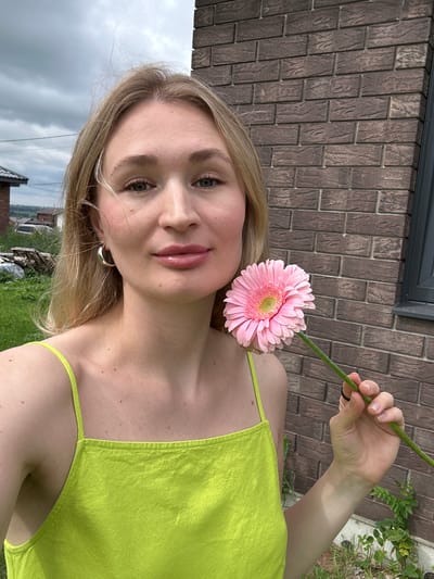 A young woman with blonde hair and green eyes, wearing a bright green tank top, holds a pink gerbera daisy in her right hand in front of a brick wall. She is outdoors, with green grass and trees in the background. The sky is overcast. The woman is looking towards the camera. The location is in the city of Mokino, Russia.
