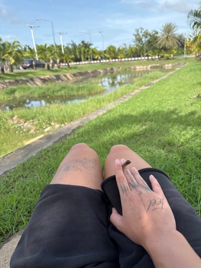 The image provides a first-person perspective of an individual seated on a grassy bank in Tambon Hua Toei, Thailand. The person wears black shorts, with a faint, light-colored tattoo visible on the upper left thigh. The right hand, featuring thin dark line tattoos across the fingers and the word "Feel" on the back, holds a partially consumed, lit cigarette or joint. In the foreground, green grass meets a concrete border alongside a narrow water channel containing grass and reflecting the sky. Beyond this, a paved road is visible, traversed by a dark sedan and lined with multiple streetlights. A dense treeline, including various palm trees, runs parallel to the road, separated by a white-and-red barrier. The sky is predominantly clear blue with scattered white clouds, indicating daytime conditions.