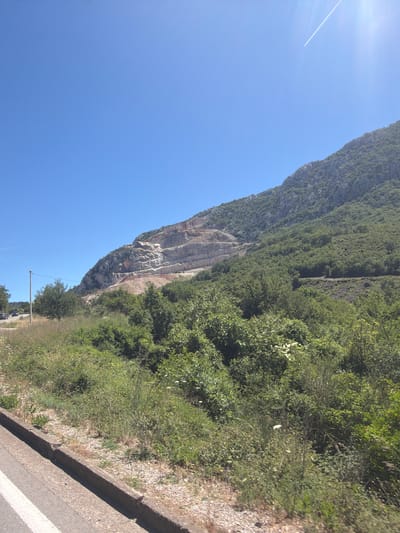 The image depicts a sunny day in Mišići, Montenegro, with a clear blue sky and a bright sun. The foreground shows a road with a white line on the left side, bordered by a concrete curb and gravel. Beyond the road, dense green vegetation, including trees and bushes, covers the lower slopes of a mountain. The mountain itself features a large, excavated quarry with distinct horizontal layers, revealing exposed rock faces. A single utility pole stands to the left of the road. In the distance, a thin white contrail stretches across the sky, indicating the presence of an airplane. The overall scene is that of a natural landscape impacted by industrial quarrying, under a bright, clear sky.