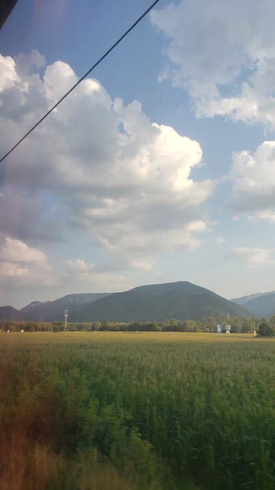 The image depicts a vast, green agricultural field, likely corn, stretching towards a line of distant, undulating, dark green mountains under a bright blue sky with scattered white, fluffy clouds. A thin, dark diagonal line, possibly a power line or cable, cuts across the upper portion of the sky. In the mid-ground, a row of trees lines the edge of the field. Several man-made structures are visible near the treeline: a tall, slender white tower with a complex structure at its apex, a bridge or overpass, and a couple of blue and white signs on poles. The overall scene suggests a rural landscape, possibly captured from a moving vehicle, given the slight motion blur in the foreground foliage. The location is identified as Uhorská Ves, Slovakia.