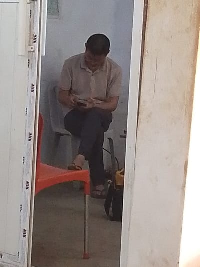 The image depicts an indoor scene in Olomoda, Nigeria, viewed through a doorway or opening. A single adult male, with short dark hair, is seated on a light-grey plastic chair, facing downwards. He wears a light-beige short-sleeved polo shirt, dark trousers, and dark open-toed sandals. His legs are crossed at the ankles, and he holds a dark-colored mobile phone in both hands, actively engaged with the device.
The immediate foreground on the left shows a partial view of an unoccupied red plastic chair with a silver-colored leg. To the right of the seated individual, on the floor, is a black backpack or bag featuring a yellow strap or accent. The walls of the room are light-colored, possibly white or light grey, and a faint, blurry image or object is visible on the wall behind the seated person. The doorframe on the left is white PVC with repetitive "AW" or "AX" markings, while the doorframe or wall section on the right appears to be a lighter, off-white, slightly textured surface.