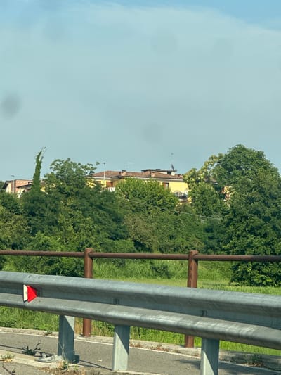 The image captures a rural or suburban scene in Pumenengo, Italy, under a clear, pale blue sky. In the foreground, a metallic guardrail runs horizontally across the frame, supported by concrete pillars. Attached to the guardrail are two red and white reflective markers, oriented towards the viewer. Beyond the guardrail, a rustic fence constructed with a dark brown rail and wooden posts separates the immediate foreground from a grassy embankment.
The middle ground is dominated by lush green foliage, dense trees, and vegetation. Peeking through the trees, on a slight incline, are sections of residential buildings. One prominent building features a yellow facade with a terracotta-tiled roof. Another building to its left is a lighter shade of brick. Antennas are visible on the rooftops. A peculiar, tall, slender tree or vine-covered structure with a pointed top stands to the left of the yellow building, intertwined with greenery.
The background consists of the pale sky, with some faint atmospheric haze. The overall impression is one of a tranquil, sunlit day in a semi-rural Italian setting. No people are visible in the frame.