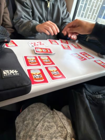 The image depicts a group of people playing a card game around a white table. The cards feature images of Duff Beer cans and some have red backgrounds with white numbers. A black bag with a "NEXT ONE" logo is visible on the left side of the table. The setting appears to be indoors, possibly on a train or in a public space, given the presence of a window with a grid pattern in the background. The people are casually dressed, and their hands are engaged in handling the cards. The lighting is natural, coming from the window. There is no indication of the city of Best, Netherlands, in the image or its surrounding context.