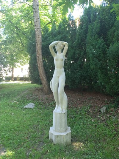 A concrete statue of a nude female figure stands on a pedestal in a grassy area with trees and dense evergreens in the background. The statue depicts a slender woman with her arms raised above her head, hands clasped behind her neck. A subtle feline or serpentine form is integrated into the sculpture, wrapping around the figure's left leg and extending upwards behind her. The statue is located in the city of Písek, Czechia. The setting appears to be a park or garden, with dappled sunlight filtering through the leaves of deciduous trees in the upper left and the dark green foliage of the evergreens forming a backdrop. The ground is covered with green grass, scattered with fallen leaves and a few small rocks. In the distance, behind the evergreens, hints of a building with windows can be seen. The scene is peaceful and natural, with the statue serving as a central focal point.