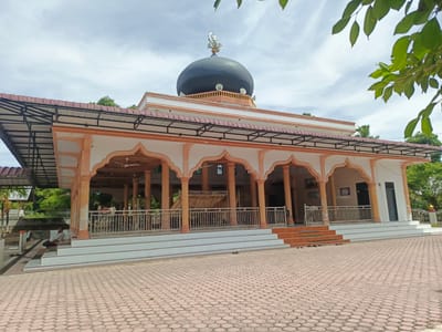 The image shows a mosque in Sawang, Indonesia, with a prominent black dome topped by a crescent moon symbol and speakers. The mosque features an open-air covered veranda with orange columns and white archways, supported by a metal roof structure. A metal railing encloses the veranda. The ground in front of the mosque is paved with hexagonal tiles. To the left of the mosque, a man is sitting on the steps, and several pairs of sandals are neatly placed on the ground. Lush green trees and foliage surround the mosque, and the sky is partly cloudy. The architectural style suggests a tropical climate.