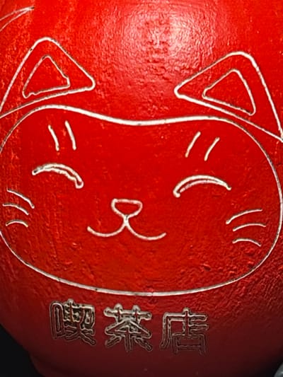 This close-up image features a red, spherical object with a stylized cat face drawn on it in white outline. The cat face has triangular ears, simple curved lines for eyes and a mouth, and short strokes for whiskers. The object appears to be a type of ornament or toy.
On the right side of the object, there are two vertical lines of text engraved in a metallic color. The top line appears to contain three characters, and the bottom line contains two characters. The characters are stylized and may be in an East Asian script.
The overall impression is of a decorative item with a playful design. The lighting suggests the photo was taken indoors, possibly in a studio setting or a well-lit room. There are no cues to suggest a specific time of day or weather conditions. The location context of Lenina, Russia, does not provide any specific visual cues within the image itself.