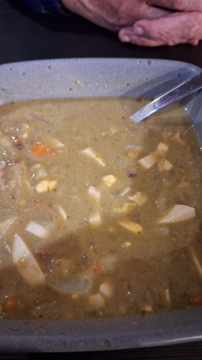 The image shows a close-up of a bowl of soup. The soup is a murky greenish-brown color and appears to contain various ingredients such as pieces of what looks like potato, carrot, and possibly egg. A silver spoon is partially visible in the upper right corner of the bowl, resting on the edge of the soup. In the background, out of focus, are two hands, suggesting someone is about to eat the soup. The overall lighting is dim. The image does not contain any information about the location in the city of Ozolnieki, Latvia.