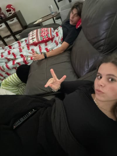 This indoor photograph, taken in Prosper, United States, features two individuals relaxing on a dark brown leather sofa. The image is oriented sideways, offering a candid perspective.

On the right, a young woman with light brown hair, wearing a black long-sleeved top, looks towards the camera with a slightly pouting yet casual expression, forming a peace sign with her right hand. A silver-toned bracelet is visible on her left wrist. She is partially covered by a black fuzzy blanket, which also conceals a black remote control tucked into its folds.

To her left, further back on the sofa, a young man with dark hair and a red bandana or head covering looks directly at the camera with a neutral expression. He wears a black t-shirt and is covered by a white blanket featuring a festive pattern of red pickup trucks and Christmas trees, suggesting a holiday theme. A red, white, and green patterned cushion with a geometric design rests on his lap. He also wears a dark watch on his left wrist.

The setting appears to be a comfortable residential living space. In the partial background on the left, a shelving unit is visible, along with a red sports helmet (possibly a football helmet) and another piece of equipment, indicating a personal touch to the room. No visible text is discernible in the image.