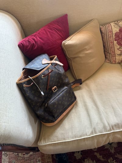 A brown Louis Vuitton backpack lies open on a light-colored, corduroy sofa. A red pillow is positioned behind the backpack, and a tan pillow and a patterned pillow with floral designs are to its right. A white charging cable protrudes from the backpack. The sofa is in a room with a patterned rug visible in the lower left corner of the image. The location is not specified as being in Chichester, United Kingdom.