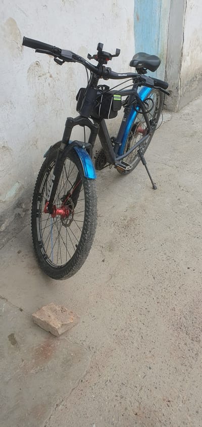 In Somoniyon, Tajikistan, a black electric bicycle with blue fenders is parked on a concrete surface against a textured, off-white wall. The bicycle has a black frame, a black seat, and black handlebars with a phone mount. It is equipped with two small black bags attached to the frame, one in front of the other, and a kickstand supporting it. The front wheel has a red hub and disc brake, and the front fender is also blue. The rear wheel is partially visible, showing spokes and a disc brake. A weathered brick lies on the ground in front of the bicycle. The environment appears to be an outdoor urban setting, possibly a street or courtyard, with a light blue painted section of the wall visible in the background.