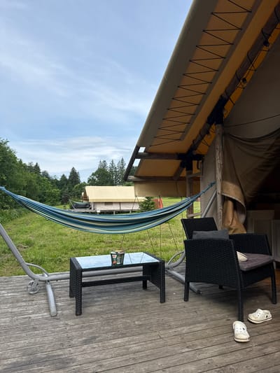 The image depicts a glamping site with a focus on outdoor relaxation. In the foreground, a weathered wooden deck is visible, with two black woven chairs, a glass-topped table, and a hammock. The table holds a cup of instant noodles and a small container. On the deck, a pair of white clogs are placed near one of the chairs.

The hammock, with blue and green stripes, is suspended from a gray metal frame, extending towards a grassy field with trees in the background. Beyond the field, a large canvas tent with a yellow roof is situated, suggesting accommodation facilities. The sky is overcast with a few wisps of clouds. The environment appears to be rural, with lush green grass and dense foliage.

This scene is located in the city of Zamosh'e (Zaklinskoe S/P), Russia, a location known for its natural beauty and outdoor recreational opportunities.