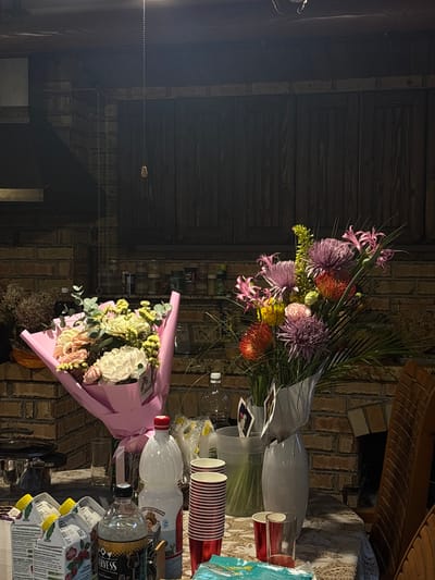The image depicts an indoor scene, possibly a kitchen or dining area, with exposed brick walls and dark wooden cabinets. A table is set with various items, including two bouquets of flowers. One bouquet, wrapped in pink paper, contains light-colored roses and other blossoms. The second bouquet is more vibrant, with purple, orange, and yellow flowers. Several beverage containers are visible on the table, including juice cartons, a bottle of water, and a bottle with a red cap. There are also stacks of red and white striped cups and some drinking glasses. In the background, shelves are stocked with jars and bottles. The lighting suggests an evening setting, with artificial light illuminating the scene. The location of Zima Yuzhnaya, Russia is not directly evident from the visual information in the image.