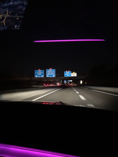 The image captures a night-time view from inside a vehicle driving on a highway, likely in or near Le Chesnay-Rocquencourt, France, as suggested by road signs. The highway is illuminated by headlights and streetlights, revealing multiple lanes and traffic. Overhead highway signs, predominantly blue with white text, indicate directions and destinations. One sign shows "ROUEN POISSY" and "ST QUENTIN EN YVELINES" with associated distances, and another shows "VERSAILLES" with a distance. A digital display, possibly part of the vehicle's navigation system, is visible in the upper left corner. A thin, bright pink illuminated strip runs horizontally across the top of the image, and another similar strip is visible along the bottom edge. The overall scene is dark, with the primary light sources being the vehicle's headlights, road lights, and the illuminated signs. The presence of other vehicles on the road indicates ongoing traffic.