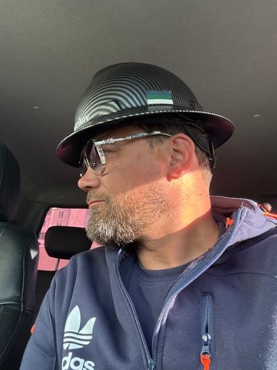 The image shows a man in a car, wearing a black carbon fiber-patterned hard hat with a small rectangular flag patch featuring green, blue, and white horizontal stripes. He is also wearing sunglasses with ornate detailing and a dark blue Adidas hooded jacket. The man has a light-colored beard and is looking to his left. Sunlight is casting a warm glow on his face and neck. The interior of the car is visible, including a black seat and the car's roof liner. The background, seen through the car window, is blurred and appears to show a building. The specific location of Usinsk, Russia, is not evident from the visual information in the image.
