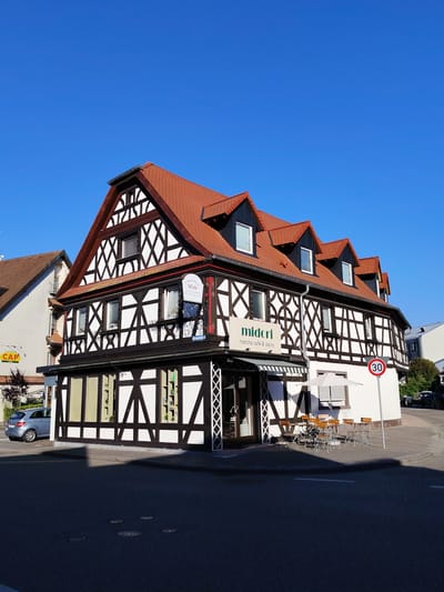 The image depicts a charming, traditional half-timbered building on a sunny day, likely in Achern, Germany. The building, with its distinctive black beams and white infill, has a prominent red-tiled roof and several dormer windows. The ground floor features large display windows and a storefront with an awning.
A sign indicates "midori matcha café & bistro," suggesting a cafe is operating within the building. Another sign, for "China-Thai Wok," is also visible, along with a phone number. Street names like "Kirchstraße" are present.
In front of the cafe, a few tables and chairs are set up under a white umbrella, implying an outdoor seating area. A speed limit sign showing "30" is positioned on the sidewalk. A silver hatchback car is parked on the street to the left of the building, and a neighboring building with a brown-tiled roof is visible to the far left.
The scene appears peaceful and quiet, with the bright sunlight casting shadows on the asphalt road in the foreground. The sky is a clear, vibrant blue, indicating fair weather and a likely daytime setting, perhaps late morning or early afternoon. The overall impression is one of a pleasant, European town street scene.