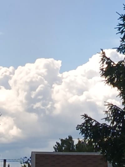 The media file captures a daytime scene dominated by a bright blue sky filled with large, puffy cumulus clouds. In the foreground, the dark green branches of a conifer tree are visible on the right, silhouetted against the sky and clouds. Below the trees and clouds, the top of a building with a brown, shingled roof and a white coping is visible. The building appears to be a house or a similar structure. In the distance, the tops of other trees can be seen, and a hint of a cloudy sky suggests a change in weather or a different layer of clouds. The overall impression is of a clear, sunny day with some dramatic cloud formations, possibly in a rural or suburban setting in Furmanov, Russia. There is no discernible text in the image. The scene evokes a sense of calm and natural beauty.