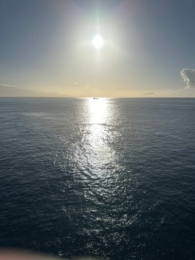 The scene depicts a wide expanse of ocean under a bright, clear sky, observed from a higher vantage point off the coast of San Sebastián de La Gomera, Spain. The sun, heavily overexposed, is positioned high in the upper-left quadrant of the sky, casting a strong, shimmering path of light directly onto the water surface, extending towards the bottom of the frame. The sky is predominantly light blue with a white haze around the sun, and a single, distinct white cloud is present in the upper-right corner. The ocean is a dark blue-grey, with its surface reflecting the sunlight in a radiant vertical band. A silhouette of a ship, possibly a cargo vessel or ferry, is visible on the horizon, directly within the sun's reflection. Faint landmasses are discernible along the horizon on both the left and right sides. No individuals are present, and no specific actions or interactions are occurring beyond the environmental elements.