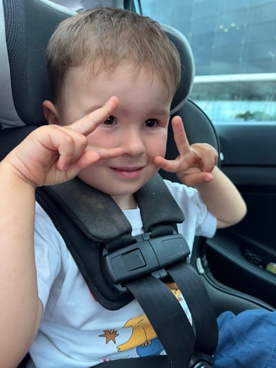 A young child, likely a boy, sits in a car seat, looking directly at the camera and making a peace sign with both hands. The child has light brown hair and is wearing a white t-shirt with a cartoon graphic and blue pants. The car seat has a black harness with a prominent buckle. The background shows the interior of a car, including a window with a blurred view of an urban environment, possibly El Prat de Llobregat, Spain, with some building structures visible. The lighting suggests it is daytime.