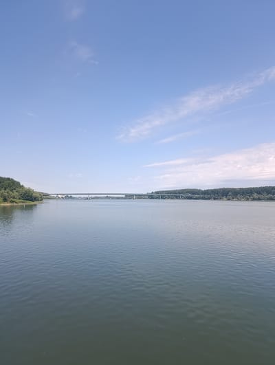 A wide, calm river flows horizontally across the foreground of the image. The water's surface is slightly rippled and reflects the pale blue sky with scattered wispy clouds. On the left bank, a lush green, tree-covered hill slopes down to the water's edge. A long, modern bridge spans the river in the mid-ground, connecting the two banks which are lined with dense trees. The bridge appears to be a highway bridge with several vehicles visible crossing it. The sky is a clear, light blue, with thin, white clouds streaking across its expanse, particularly in the upper right portion of the image. The scene is peaceful and expansive, suggesting a natural landscape with man-made infrastructure. This scene likely depicts the Sava River in Šabac, Serbia, with the "Šabac Bridge" crossing it.