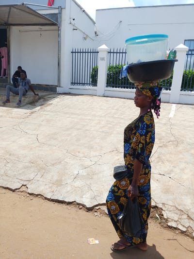 The image shows a woman carrying a large basin filled with items on her head, walking on a cracked concrete ground. She is wearing a colorful floral dress and a headscarf. A black bag is slung over her shoulder, and she holds another black bag in her right hand. Her hair is styled in braids with pink accents.

In the background, two men are sitting on steps in front of a white building with a covered entrance. One man is wearing a grey shirt and pants, while the other is wearing a black shirt and grey pants. Further back, a black metal fence encloses an area with green bushes. A few security cameras are visible on the white building. The scene appears to be outdoors during the daytime, under a bright sky. The presence of the woman carrying goods on her head suggests a common practice for vendors or people transporting items in this region. The cracked pavement and the style of the building hint at a location in Iseyin, Nigeria. There is no discernible text in the image, except for a small, illegible sticker on the ground in the lower right corner.