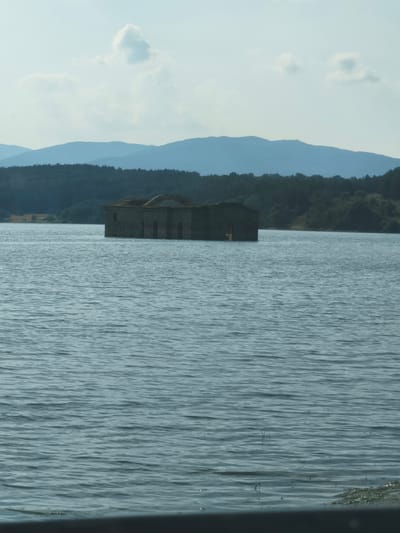 The media file depicts a submerged stone building, likely a church, standing partially in the middle of a calm body of water. The building has a distinct arched roof and visible windows and doorways, though they are partially obscured by the water. The water's surface reflects the soft light of the sky, creating shimmering patterns. In the background, a dense forest covers rolling hills, which in turn are backed by distant, hazy blue mountains. The sky is a pale blue, with a few scattered clouds. The overall scene suggests a serene and somewhat melancholic atmosphere, hinting at a past event that led to the building's inundation. The time of day appears to be afternoon or late morning, with diffused sunlight. The weather seems pleasant and clear. There is no visible text in the image. The location is identified as Orizari, Bulgaria, which provides context for the type of architecture and landscape.