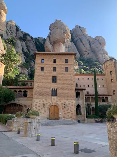 The image shows a view of the Montserrat Monastery in Monistrol de Montserrat, Spain. The monastery is built into the side of a large, rocky mountain range. The architecture of the monastery is characterized by its use of stone, with multi-story buildings featuring arched walkways and numerous windows. In the foreground, there is a large paved courtyard with several bollards. On the right side of the courtyard, a few people are seated on a stone bench, and a tall, slender cypress tree stands against the monastery buildings. The sky is clear and blue.